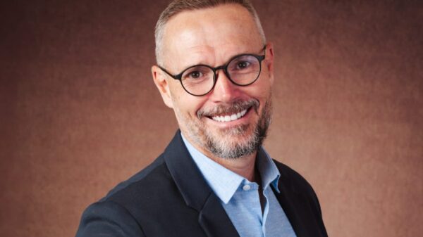 Retrato de meio corpo de um homem de terno escuro e camisa azul, com mão tatuada à frente do peito, em fundo neutro marrom, em pose profissional.