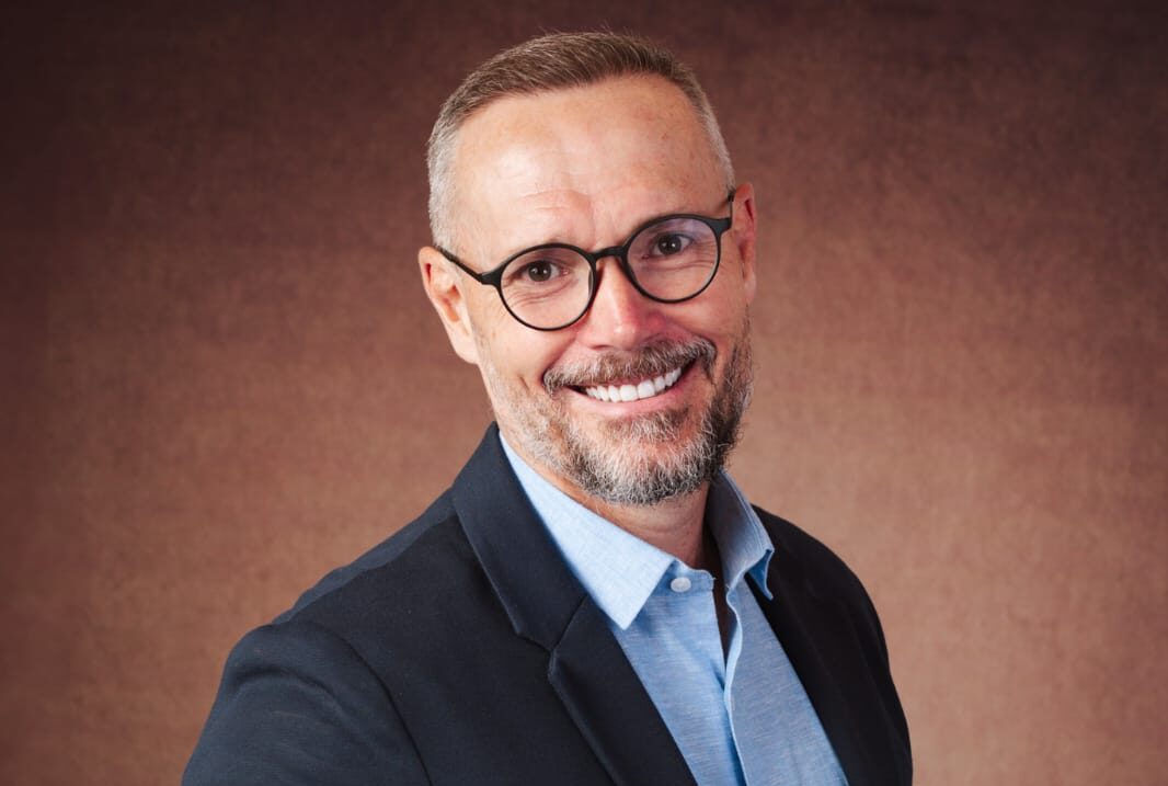 Retrato de meio corpo de um homem de terno escuro e camisa azul, com mão tatuada à frente do peito, em fundo neutro marrom, em pose profissional.